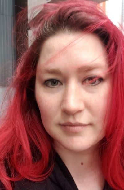Close up image of a white woman with dyed red hair, green eyes, and nose ring. Wearing a black coat.
